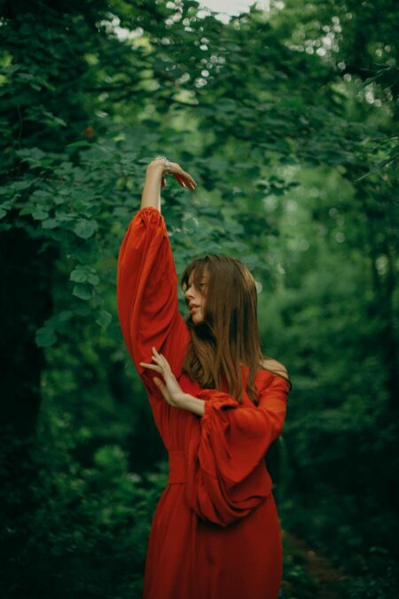Žena u crvenoj haljini s dugim rukavima pleše u šumi, okružena zelenim lišćem.