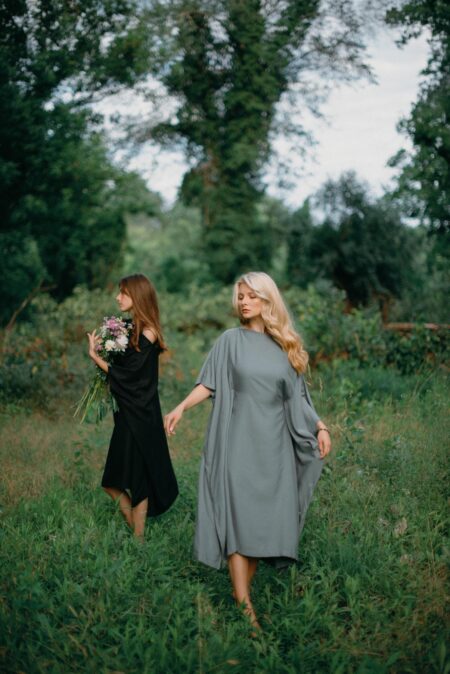 Dve žene u prirodi, jedna nosi crnu, a druga sivu kaftan haljinu, stojeći među drvećem i zelenilom.